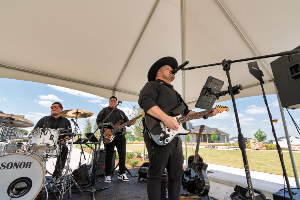 The Louis Adams Symphony performing under a tent at the Austin Point Open House Party with electric guitar, drums, and bass.