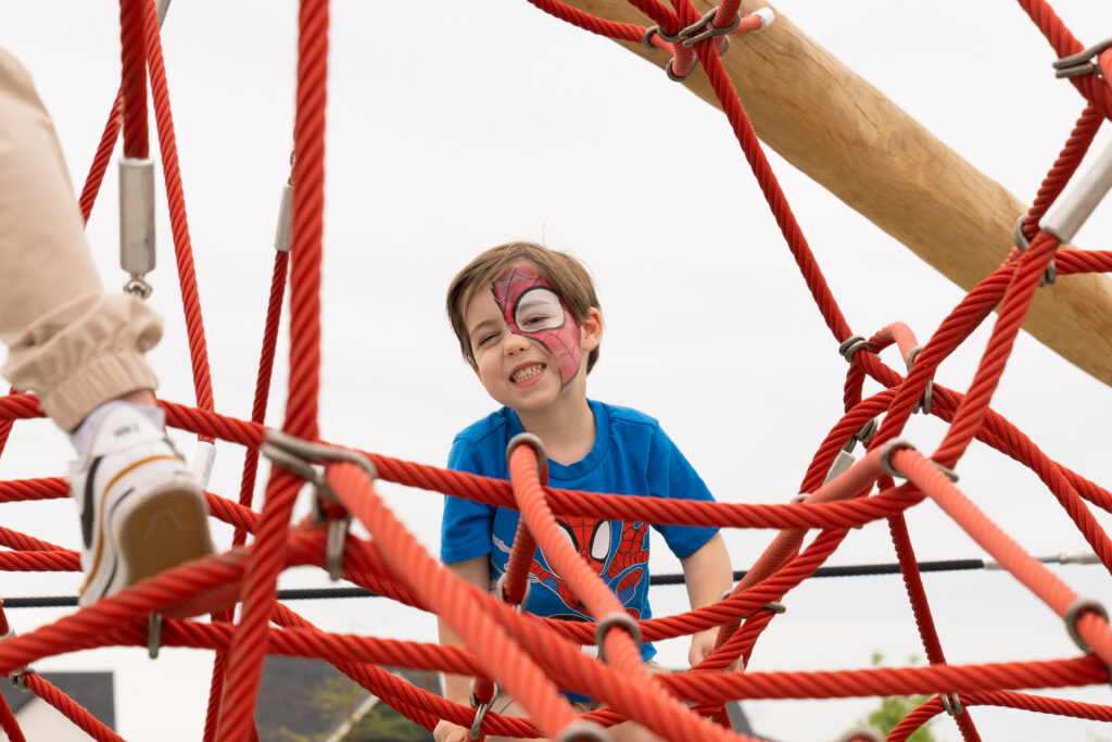 Child with face paint climbing the playground rope structure at Austin Point’s civic green.